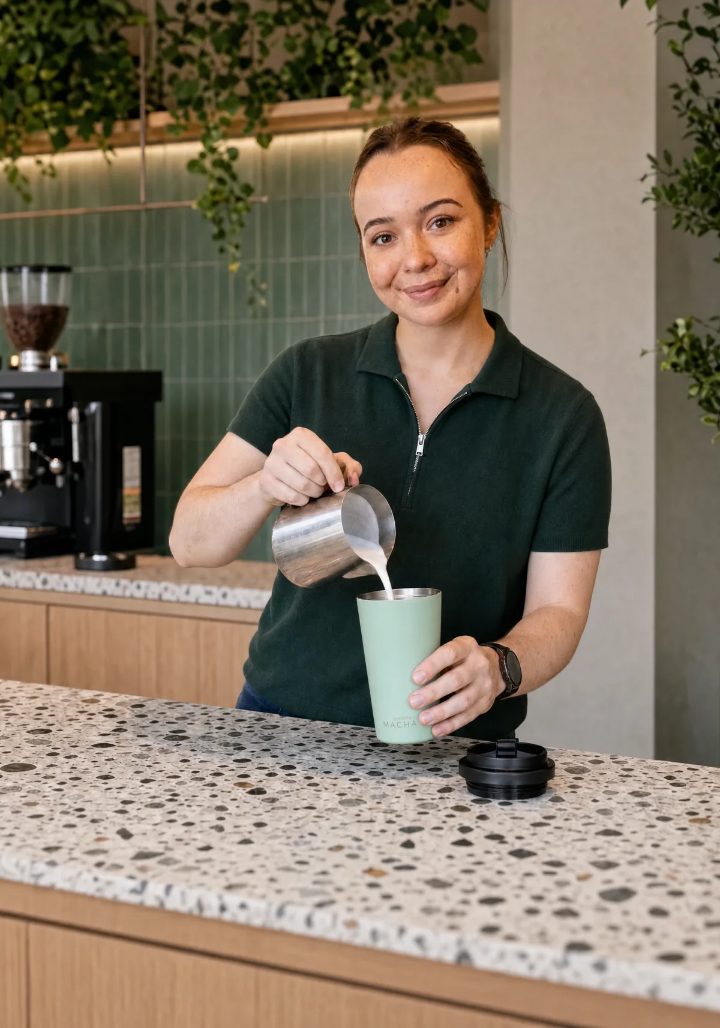 A barista pouring steamed milk from a jug into the MACHA Cup in Pistacchio, its lid resting on the counter