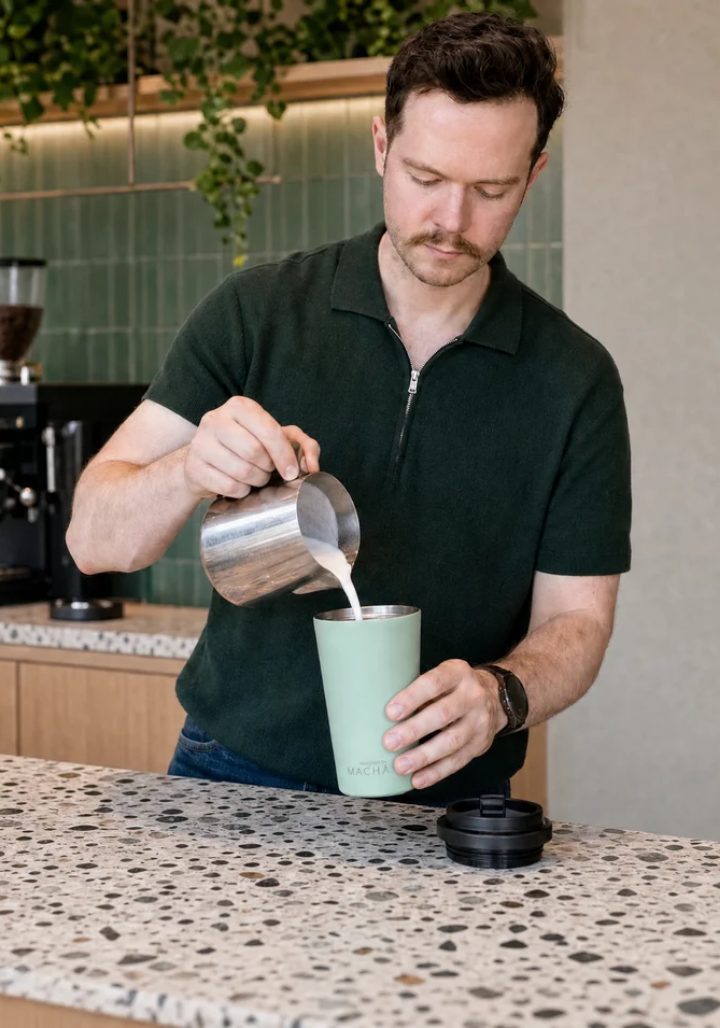 A barista pouring steamed milk from a jug into the MACHA Cup in Pistacchio, its lid resting on the counter