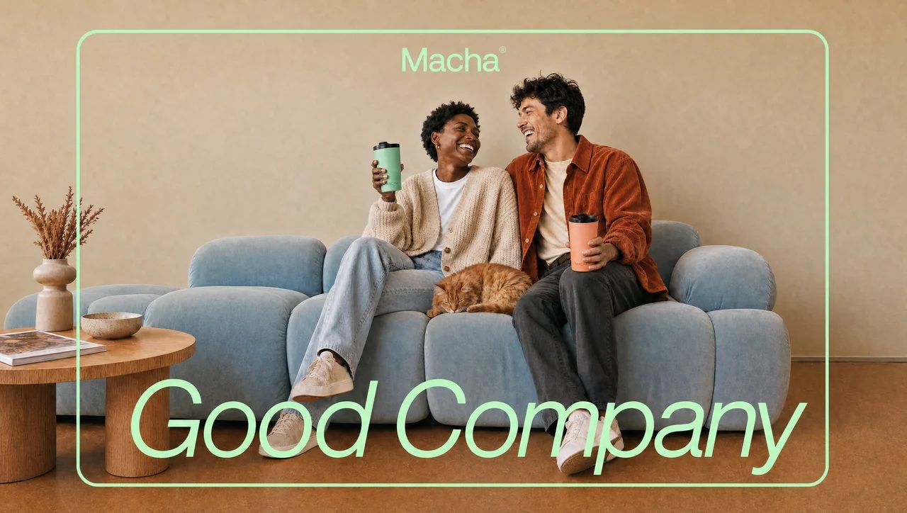 Two people laughing on a sofa with a cat between them, each holding a MACHA Cup. The words "good company" are set across the photograph in mint italic script, with the Macha wordmark above.