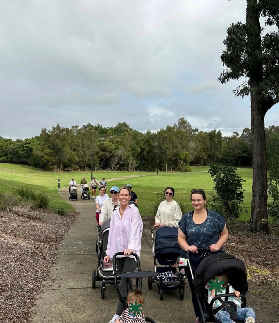 Mothers walking in single file along a park path with their prams, more of them further back up the hill